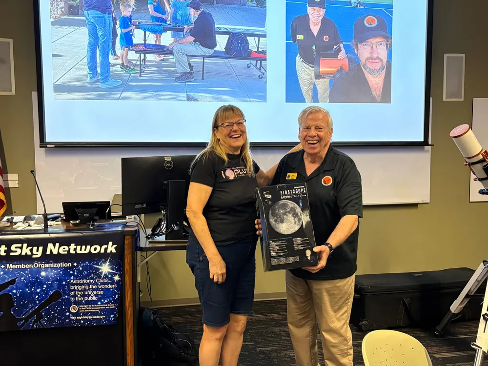 A PAS officer presenting another member with a FirstScope Moon telescope, with outreach event photos on screen behind them